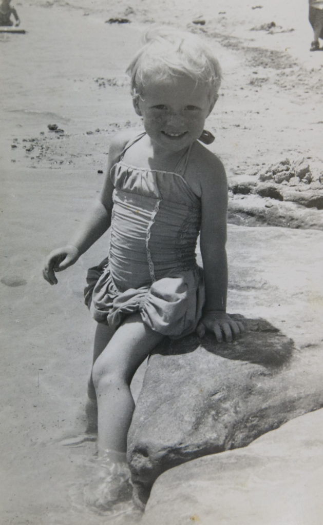 A black and white photograph of a two year old girl standing in shallow water wearing a swimming costume made with elasticised shirring.